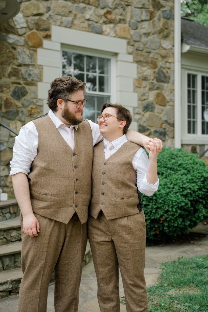 Two white queer men stand together in match brown tweed suits. One is taller and bearded.