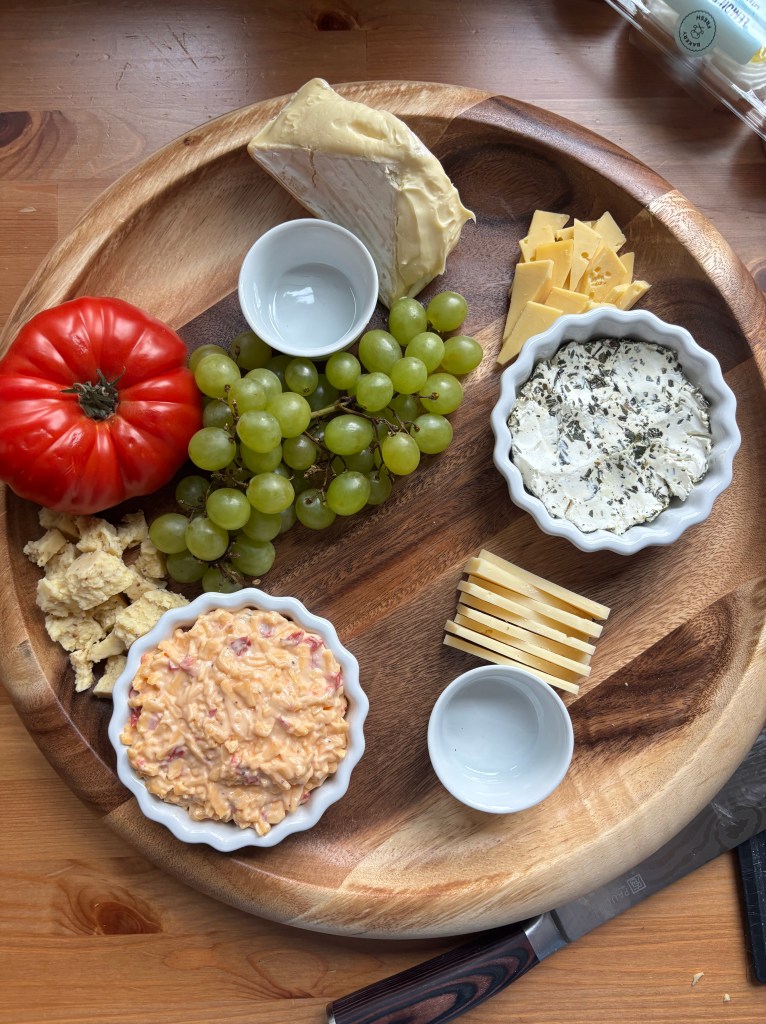 A cheeseboard with the cheese in place, plus grapes and a large tomato in place. The rest of the board is empty.