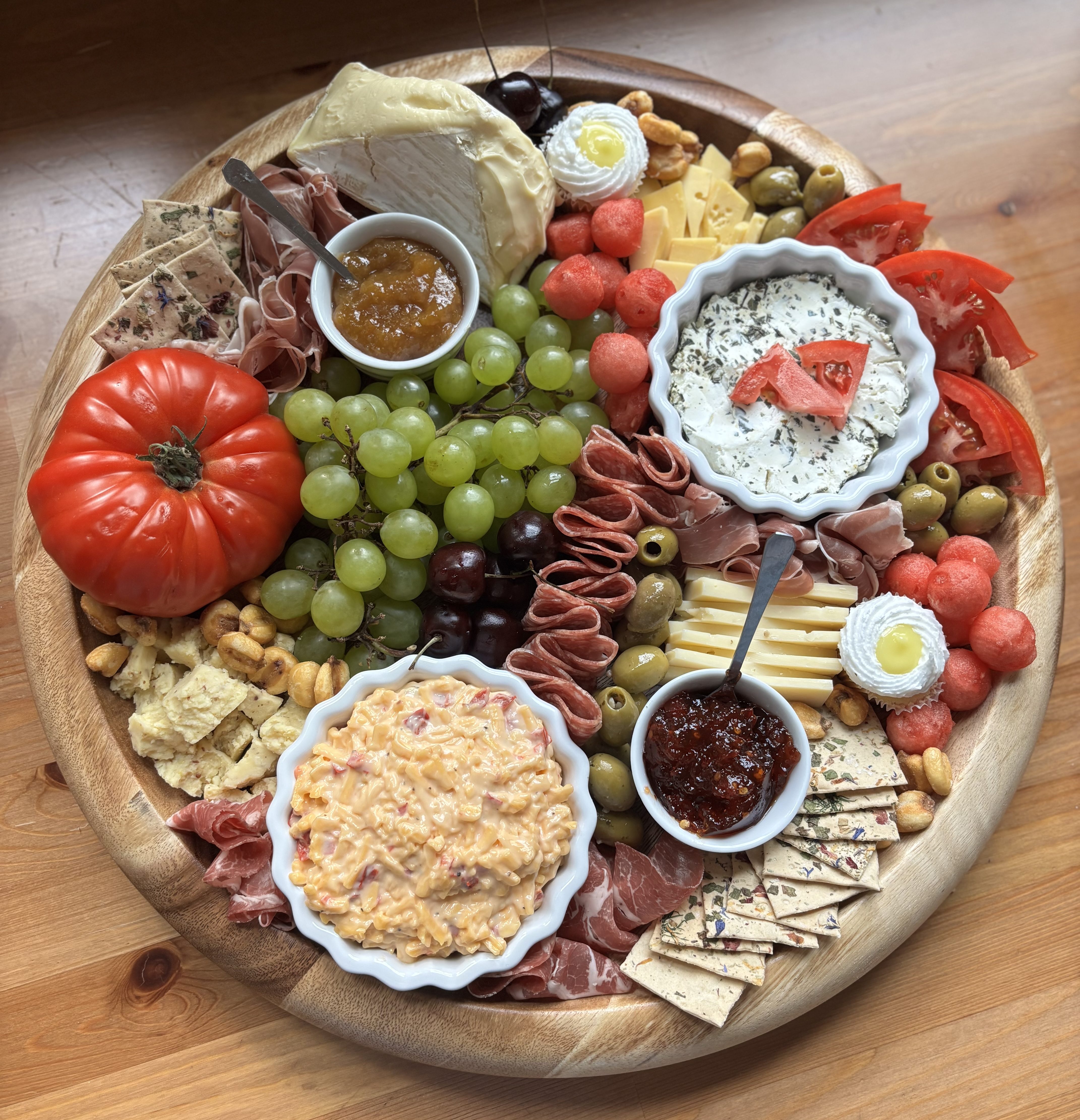 A charcuterie board. The board has tomatoes, grapes, cherries, watermelon, and a bowl of pimento cheese in addition to the typical meat and cheese.